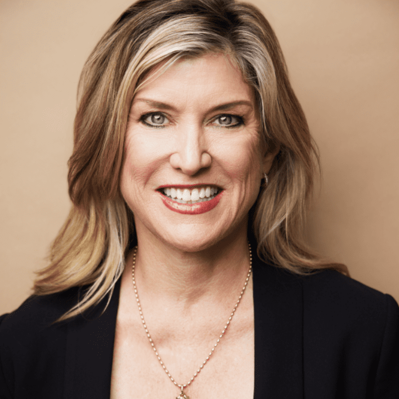A woman with shoulder-length blonde hair, wearing a black blazer and gold necklace, smiles against a plain beige background, captured in a portrait by Mike Pine.