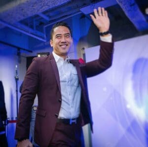 Jeremy Wong, in a maroon suit, waves and smiles while walking indoors at a formal event.