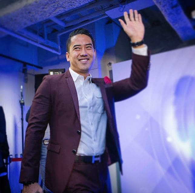 Jeremy Wong, in a maroon suit, waves and smiles while walking indoors at a formal event.
