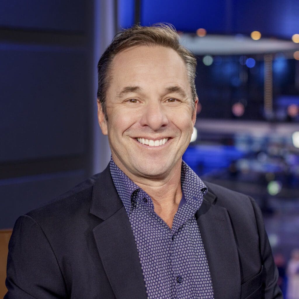 Mike Pine, wearing a dark blazer and patterned shirt, smiles at the camera in a modern, brightly lit indoor setting.