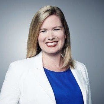 Paula Allegretti, with straight blonde hair, wears a white blazer over a blue top and smiles at the camera against a plain gray background.