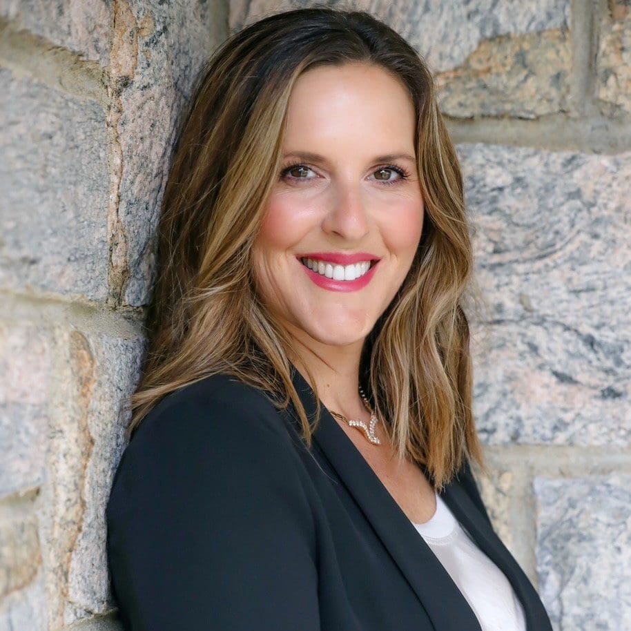 A woman with shoulder-length brown hair wearing a black blazer and white top smiles while standing against a stone wall, photographed by Mike Pine.