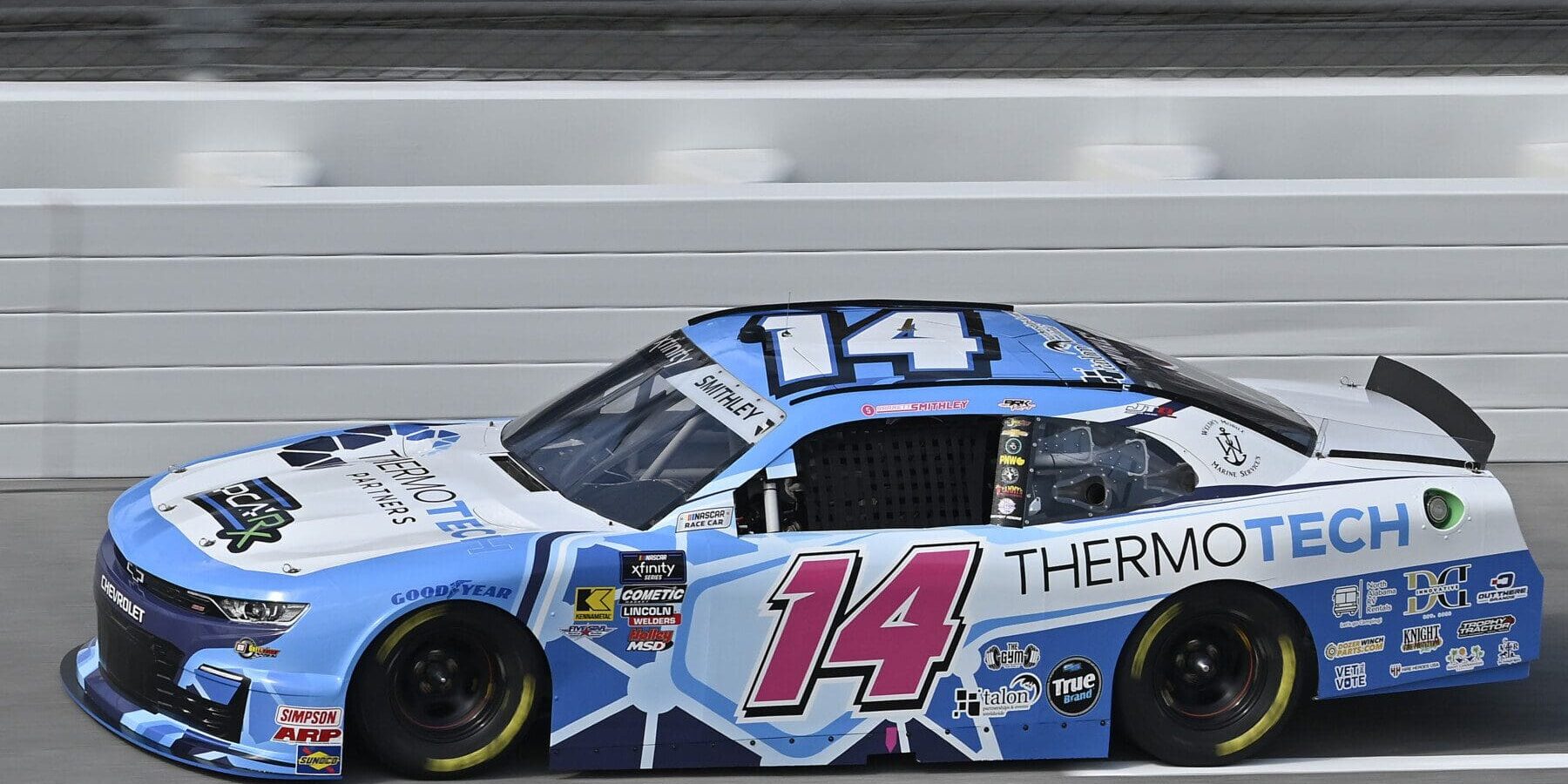 A blue and white race car with the number 14 and ThermoTech branding, promoted by a leading sports and entertainment marketing agency, speeds along a racetrack next to a white barrier.