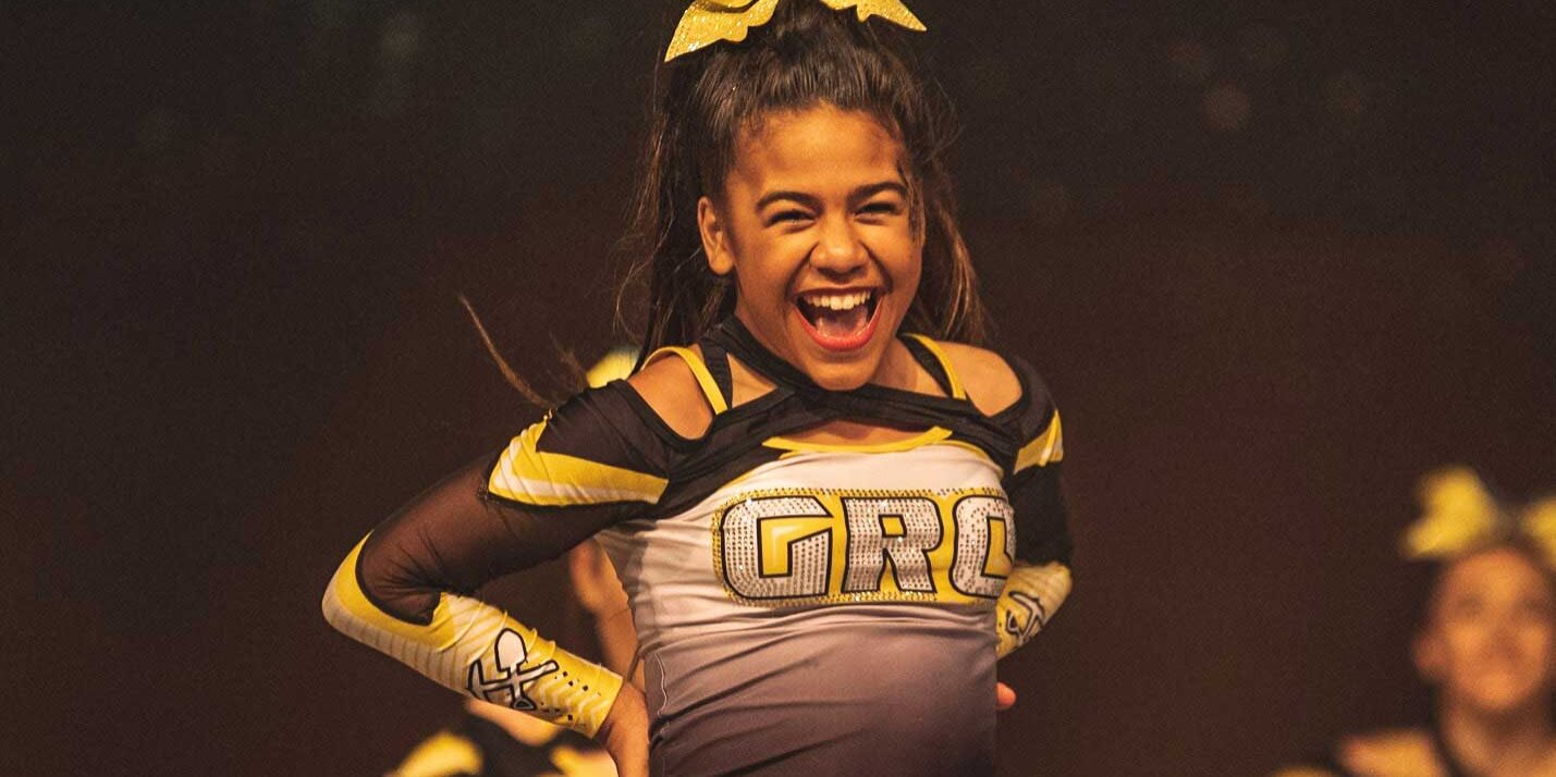 A young cheerleader in a black and yellow uniform, representing a global sports and entertainment agency, smiles with hands on hips during a performance.