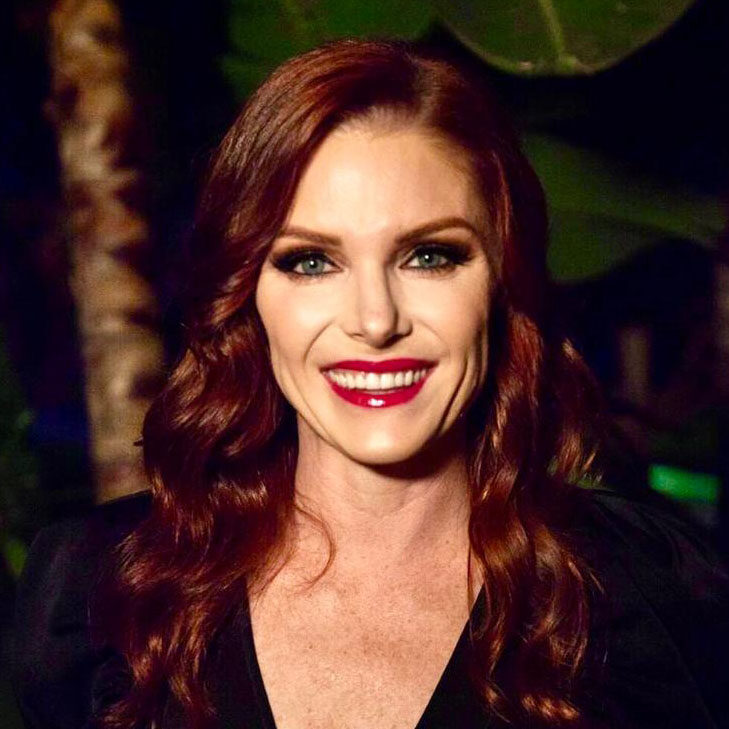 A woman with long wavy red hair, fair skin, and bright red lipstick is smiling at the camera against a dark, leafy background in a portrait by Mike Pine.