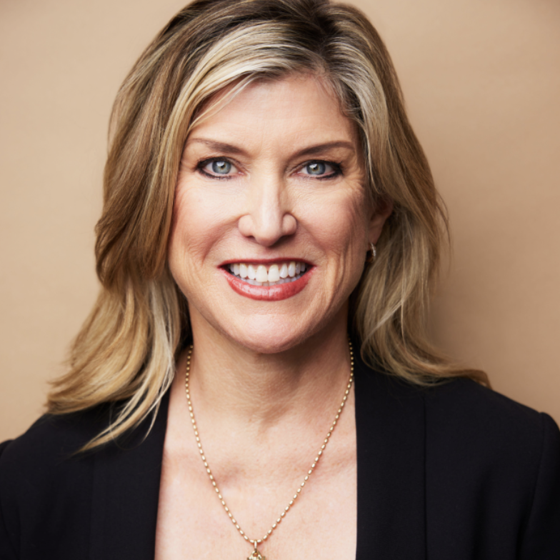 A woman with shoulder-length blonde hair, wearing a black blazer and gold necklace, smiles against a plain beige background, captured in a portrait by Mike Pine.