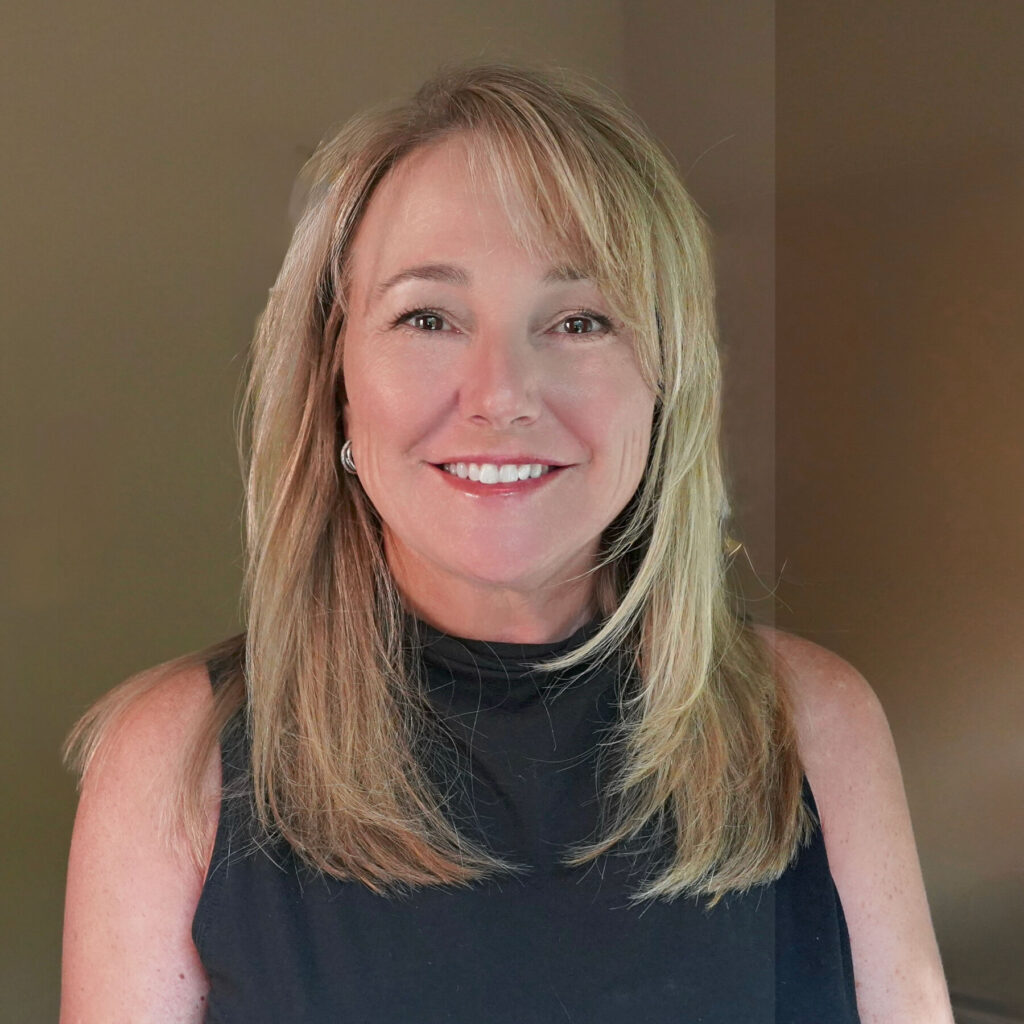 A woman with shoulder-length blonde hair wearing a sleeveless black top smiles at the camera against a plain background, photographed by Mike Pine.