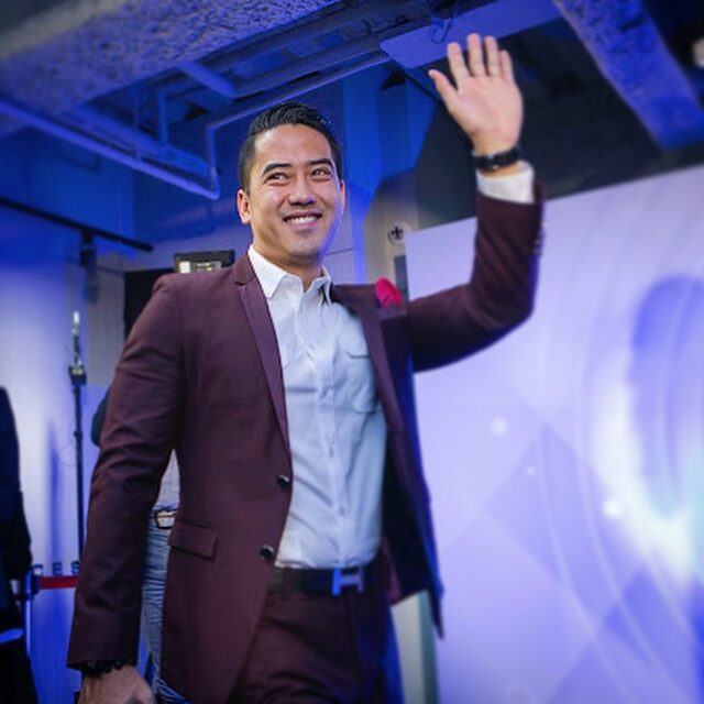 Jeremy Wong, in a maroon suit, waves and smiles while walking indoors at a formal event.
