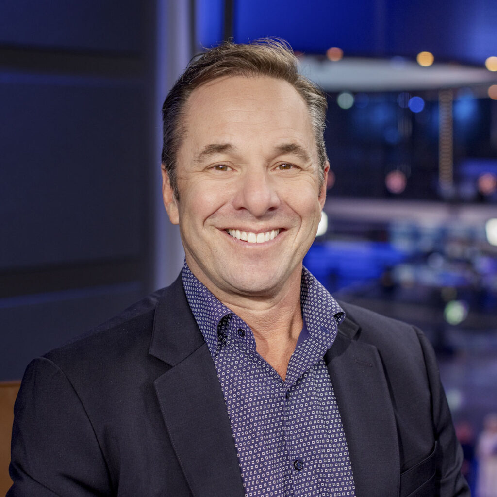 Mike Pine, wearing a dark blazer and patterned shirt, smiles at the camera in a modern, brightly lit indoor setting.
