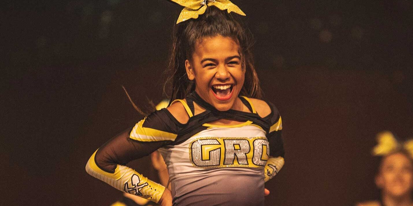 A cheerleader in a black and gold uniform smiles with hands on hips during a performance, creating an energetic moment perfect for a sports and entertainment marketing agency.