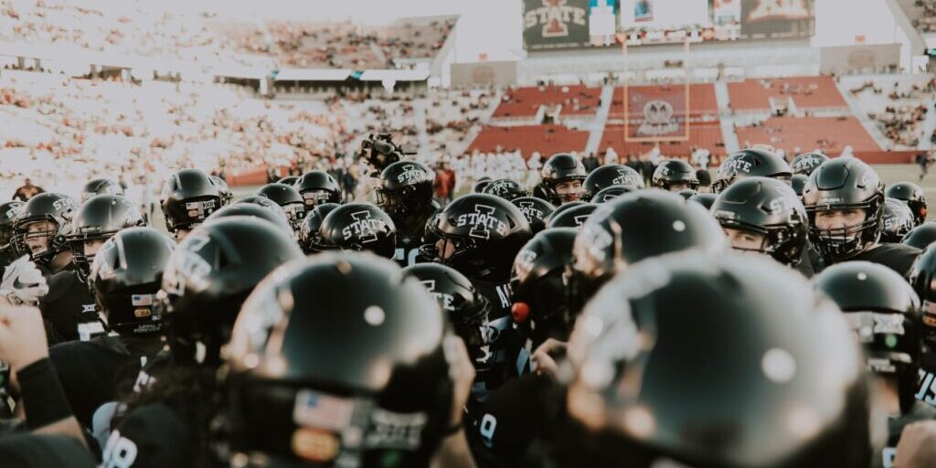 A group of football players in black helmets huddle on the field, with a stadium—buzzing from fans and empty red seats—capturing the energy often celebrated by a global sports and entertainment agency.