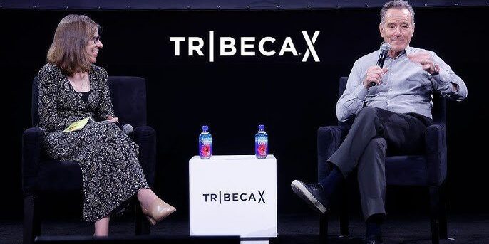 Two people sit on stage in armchairs having a conversation, with TribecaX branding behind them and two water bottles on a small table between them.