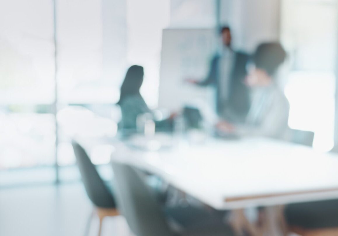 Blurry image of people sitting at a conference table, with one person standing and presenting near a whiteboard in a bright office setting at a global sports and entertainment agency.