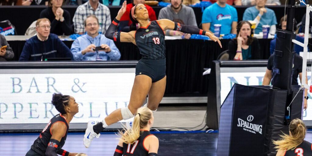 A volleyball player in black uniform leaps to spike the ball during a match, while teammates and spectators watch intently.