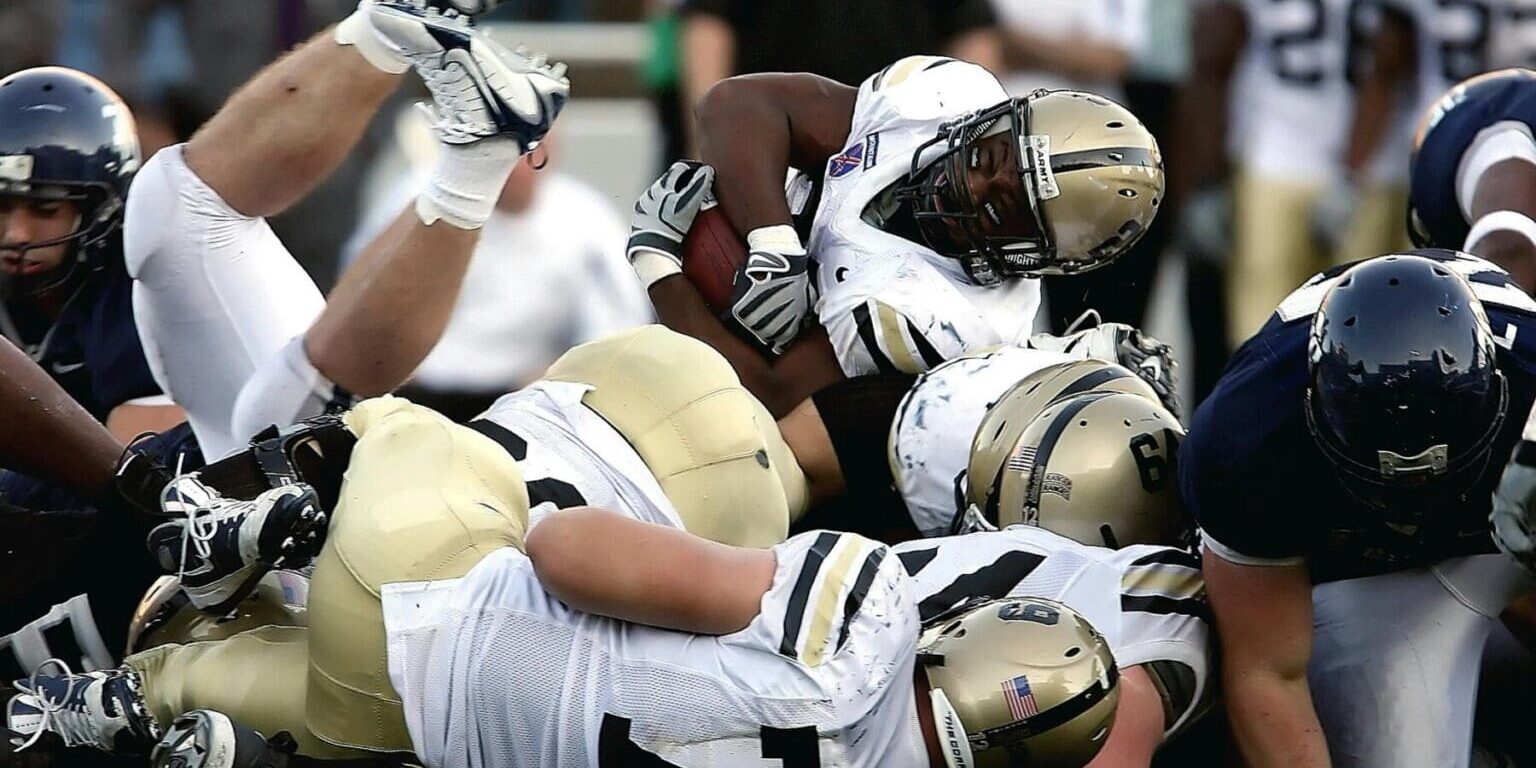 Football players from two teams collide and pile up on the field during a tackle, as the player holding the ball is pushed to the ground in an intense moment captured by a global sports and entertainment agency.